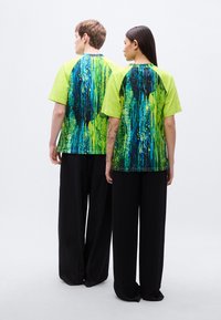 Κοντομάνικο πουκάμισο σε νέον πράσινο βαμβάκι με υφασμένο αφηρημένο μπλε και κίτρινο μοτίβο στην πλάτη· συνδυασμένο με μαύρο φαρδύ παντελόνι.