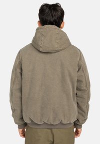 Veste à capuche vert olive avec une finition texturée, capuche ajustable et poignets côtelés, mettant en avant des coutures dessinées et un ourlet cintré.