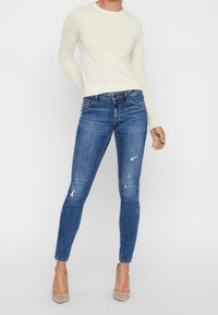 Ljus krämfärgad långärmad topp tillsammans med mörkblå skinny jeans med slitna detaljer, kompletterat med beigefärgade högklackade skor.