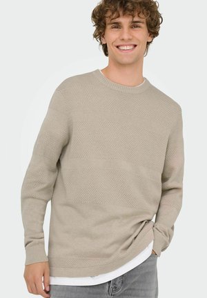 Hellbeige gestrickter Pullover mit strukturierten Streifen, geripptem Ausschnitt und Bündchen, mit einem lockeren Schnitt über einem weißen, geschichteten Hemd.