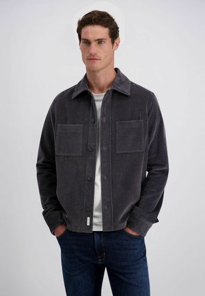 Mann mit kurzen braunen Haaren, der eine dunkelgraue Cordjacke mit Knöpfen über einem weißen Hemd und blauen Jeans trägt, die Hände in den Taschen.