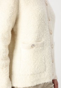 Krämfärgad cardigan i mjukt, strukturerat tyg med en stickad ficka och dekorativa knappar längs fram.