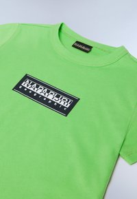 Zaļš kokvilnas krekls ar apaļu kaklu, kura priekšā ir melns taisnstūra logo ar tekstu "NAPAPIJRI geographic."
