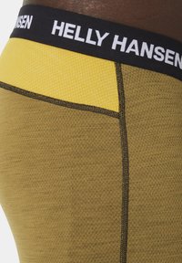 Termální legíny Helly Hansen s černým elastickým pasem, bílým logem, žlutou síťovanou vložkou a texturovanou olivově zelenou tkaninou.