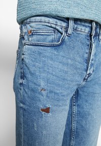 Jeans de denim azul claro con corte ajustado, que presentan parches desgastados y un sutil desvanecimiento. Diseño clásico de cinco bolsillos con cierre de botón.