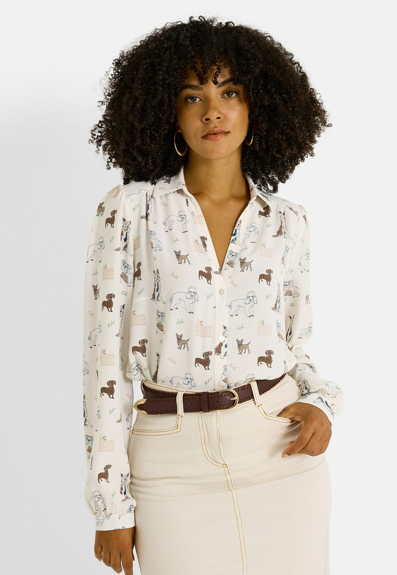Chemise blanche boutonnée avec un motif de chiens multicolores, manches longues, col et épaules froncées. Portée avec une ceinture marron.