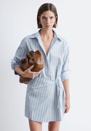 Kobieta w jasnoniebieskiej sukience w paski, trzymająca dużą brązową skórzaną torbę na jednolitym białym tle.