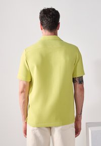 Camisa de manga corta en una tela verde claro texturizada, con collar puntiagudo y detalles de costura visibles; combinada con pantalones de color claro.