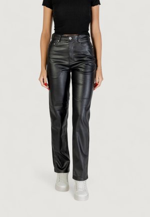 Donna che indossa un top nero a maniche corte, pantaloni in pelle nera a vita alta, sneakers bianche e un braccialetto d'argento, in piedi su uno sfondo bianco.