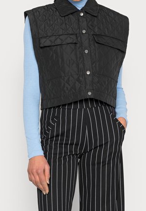 Gilet nero trapuntato senza maniche con due tasche sul petto, chiusure frontali a scatto e design accorciato, abbinato a una camicia a maniche lunghe blu e pantaloni a righe.
