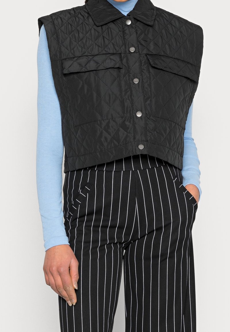Gilet nero trapuntato senza maniche con due tasche sul petto, chiusure frontali a scatto e design accorciato, abbinato a una camicia a maniche lunghe blu e pantaloni a righe.