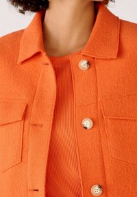 Orange ulljacka med krage, dubbla bröstfickor och två vita knappar; bärs över en ribbad orange topp.