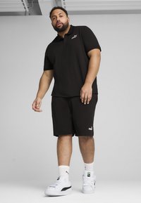 Čierne polo tričko a šortky vyrobené z tkaného materiálu, s malým bielym logom Puma na hrudi a ľavej nohe. Biele tenisky s čiernymi akcentmi.