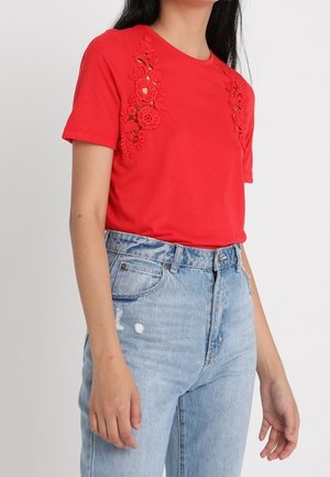 Woman wearing a red short-sleeve shirt with floral embroidery and high-waisted light blue denim jeans, standing with arms relaxed.