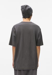 Pelēks, pārāk liels T-krekls ar īsām piedurknēm, apaļu dekoltē un mīkstu tekstūru. Valkāts ar atbilstošu pelēku cepuri, skatīts no mugurpuses.