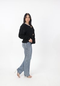 Cardigan nero e soffice con bottoni dorati, abbinato a jeans a gamba dritta azzurri e sandali con tacco trasparente. Sfondo bianco uniforme.