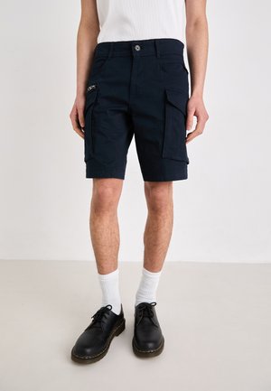 Shorts cargo navy avec plusieurs poches, une poche latérale zippée et une fermeture à bouton, associés à des chaussures noires et des chaussettes blanches.