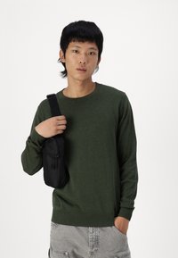 Grønn strikket genser med ribbestrikkede mansjetter, kombinert med grå shorts. En liten svart crossover-veske med glidelåser bæres av brukeren.