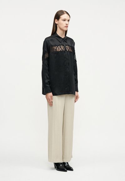 Γυναίκα που φοράει μαύρο σατέν μπλούζα με δαντελένιες λεπτομέρειες, συνδυασμένη με μπεζ παντελόνι φαρδιάς γραμμής και μαύρα παπούτσια με μυτερή μύτη, στέκεται μπροστά σε λευκό φόντο.