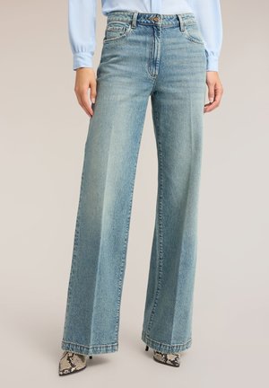 Vrouw die lichtblauwe jeans met wijde pijpen, lichtblauw overhemd en puntige hoge hakken met slangprint draagt, staat met handen ontspannen langs haar zij.