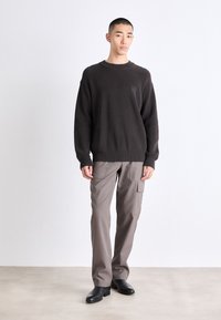 Maglione marrone a coste con collo rotondo, abbinato a pantaloni cargo grigio chiaro con tasche. Il modello indossa stivali neri, in piedi su un pavimento neutro.
