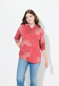 Haut à capuche rouge avec fermeture éclair sur le devant, orné de motifs en rayons de soleil blancs. Manches longues et coupe décontractée, assorti à un jean bleu clair. Texture lisse.