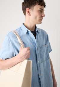 Camisa de mezclilla azul claro de botones con mangas cortas, que presenta un bolsillo en el pecho. Sosteniendo una bolsa de lona beige sencilla.