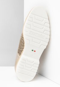 Semelle en caoutchouc blanche d'une chaussure beige avec le logo NeroGiardini, le drapeau italien et la taille 37, placée en angle contre un mur blanc d'angle.