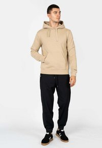 Felpa beige con tasca frontale e cappuccio con cordino, abbinata a pantaloni jogger neri con polsini elasticizzati e tasche laterali. Sneakers nere.