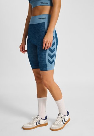 Frau trägt blaue nahtlose Sportshorts und passendes Crop-Top mit weißen Sneakern und hohen weißen Socken, steht auf grauem Hintergrund.
