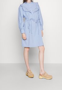 Ljusblå klänning med krage, detaljerad volangkrage, långa ärmar och en åtstramad midja. Modellen har på sig beige träskor.