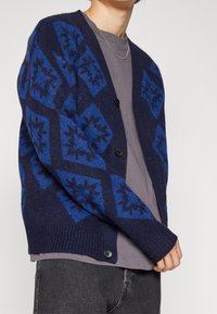 Marinblå cardigan med diamantmönster i blått och svart. Stickad tyg, knäppning med knappar och ribbade ärmslut. Bärs över en grå T-shirt.