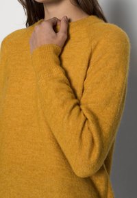 Frau in einem senfgelben Wollpullover, die mit einer Hand den Ausschnitt berührt, vor einem schlichten grauen Hintergrund.