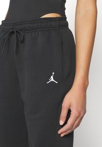 Närbild av en person som bär svarta sweatpants med en vit Jumpman-logotyp nära fickan och en svart topp.
