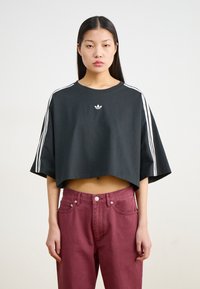 Melna  cropped t-kreklā ar baltām svītrām plecu daļā un mazu logo uz krūtīm, kopā ar bordo augstiem džinsiem.