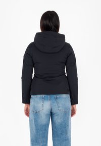 Giacca nera con cappuccio dal design elegante e tessuto testurizzato, abbinata a jeans blu chiaro. Presenti dettagli di cuciture e accenti.
