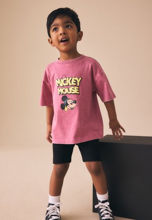 Mladý chlapec stojaci pri čiernom bloku má na sebe ružové tričko s Mickey Mousom, čierne šortky, biele ponožky a čierne tenisky.
