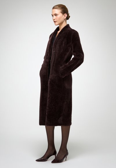 Brūns mākslīgās kažokādas mētelis ar garām piedurknēm, atvērtu priekšu, sānu kabatām un ceļgalu garuma griezumu. Valkāts pār melnu apģērbu un saskaņots ar spīdīgām kurpēm uz papēžiem.