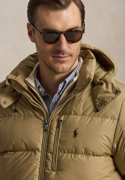 Man wearing dark sunglasses, a tan puffer jacket with a hood, and a striped collared shirt beneath, looking slightly to the side.