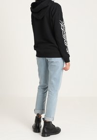 Sudadera negra con capucha y gráfico de llamas blancas en la manga, combinada con jeans azul claro con puños y botas negras hasta el tobillo.