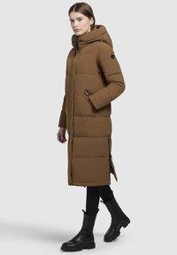 Nő, aki hosszú barna steppelt kabátot visel kapucnival, fekete leggingssel és fekete bokacsizmával, egy sima háttér előtt áll.