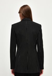 Nainen, jolla on suorat ruskeat hiukset, käyttää mustaa bleiseriä, jossa on valkoisia pystysuoria raitoja, nähtynä takaa yksinkertaista taustaa vasten.