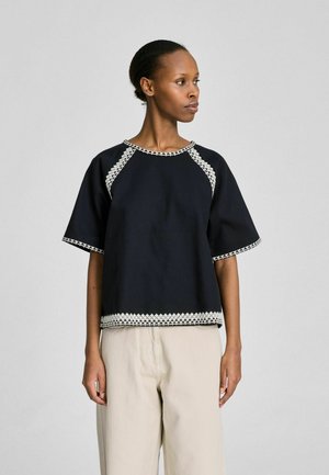 Woman wearing a black short-sleeve top with white geometric trim and beige pants, looking to her left against a plain light gray background.