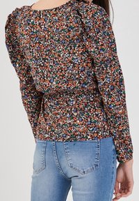 Blouse fleurie à manches longues bouffantes, avec un fond noir et des motifs floraux multicolores ; associée à un jean bleu clair.