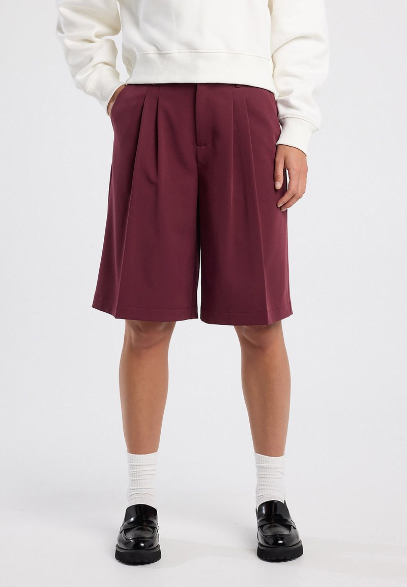 Burgundy lyhyt hame, joka on valmistettu sileästä kankaasta, yhdistetty valkoisiin ryppyisiin sukkiin ja mustiin loafer-kengiin.