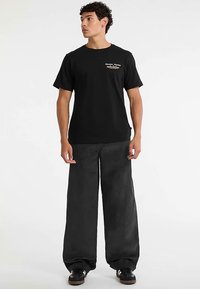 Maglietta nera in cotone con una piccola grafica e maniche corte. Abbinata a pantaloni grigi a gamba larga realizzati in materiale testurizzato.