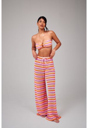 Vrouw staat blootsvoets en draagt een kleurrijk bandeau-top met zigzagpatroon en bijpassende wijdvallende broek tegen een effen grijze achtergrond.
