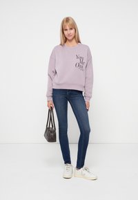 Fioletowa bluza z napisem "You Me Out", w połączeniu z ciemnoniebieskimi obcisłymi dżinsami i białymi sneakersami. Modelka trzyma małą szaro-skalną torebkę.