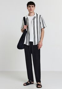 Giovane uomo che indossa una camicia a maniche corte bianca e nera a righe, pantaloni neri, sandali e porta una borsa a tracolla nera su sfondo neutro.