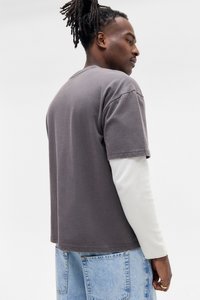 Mand med dreadlocks har en mørkegrå kortærmet skjorte lagd over en hvid langærmet skjorte og lyseblå jeans, står på siden.